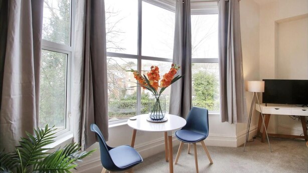 Una habitación con un ventanal grande, una mesa redonda con dos sillas, un jarrón con flores y un televisor sobre un mueble bajo.