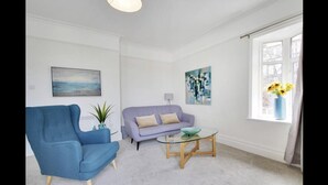 A living room with a blue armchair, a light purple sofa, and a glass coffee table. There are two paintings on the walls, a window with a view, and a vase with flowers on the windowsill. - Stunning 2-bed Apartment in Tunbridge Wells (Royal Tunbridge Wells)