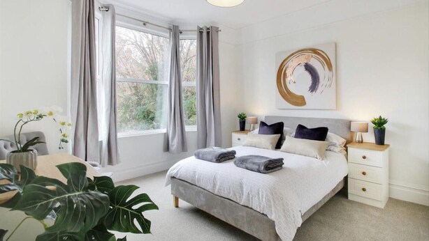 A modern bedroom with a large bed, bedside tables, a window with curtains, and a painting on the wall. - Stunning 2-bed Apartment in Tunbridge Wells (Royal Tunbridge Wells)