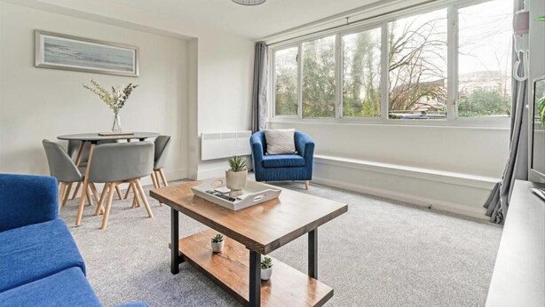 Un salon moderne avec un canapé bleu, une table basse et un coin repas comprenant une table et des chaises. Une grande fenêtre offre une vue sur les arbres et un bâtiment à l’extérieur.