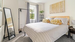 Une chambre avec un grand lit, un grand miroir, un radiateur, une fenêtre avec des rideaux, une plante et une œuvre d’art accrochée au mur.