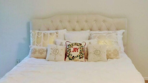 Un lit bien fait, avec une couette blanche, plusieurs coussins décoratifs et un coussin central orné du mot « JOIE ».