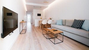 Una sala de estar moderna con un televisor de pantalla plana, una mesa de centro de madera, un sofá con cojines estampados y una zona de comedor con sillas y una pequeña mesa.