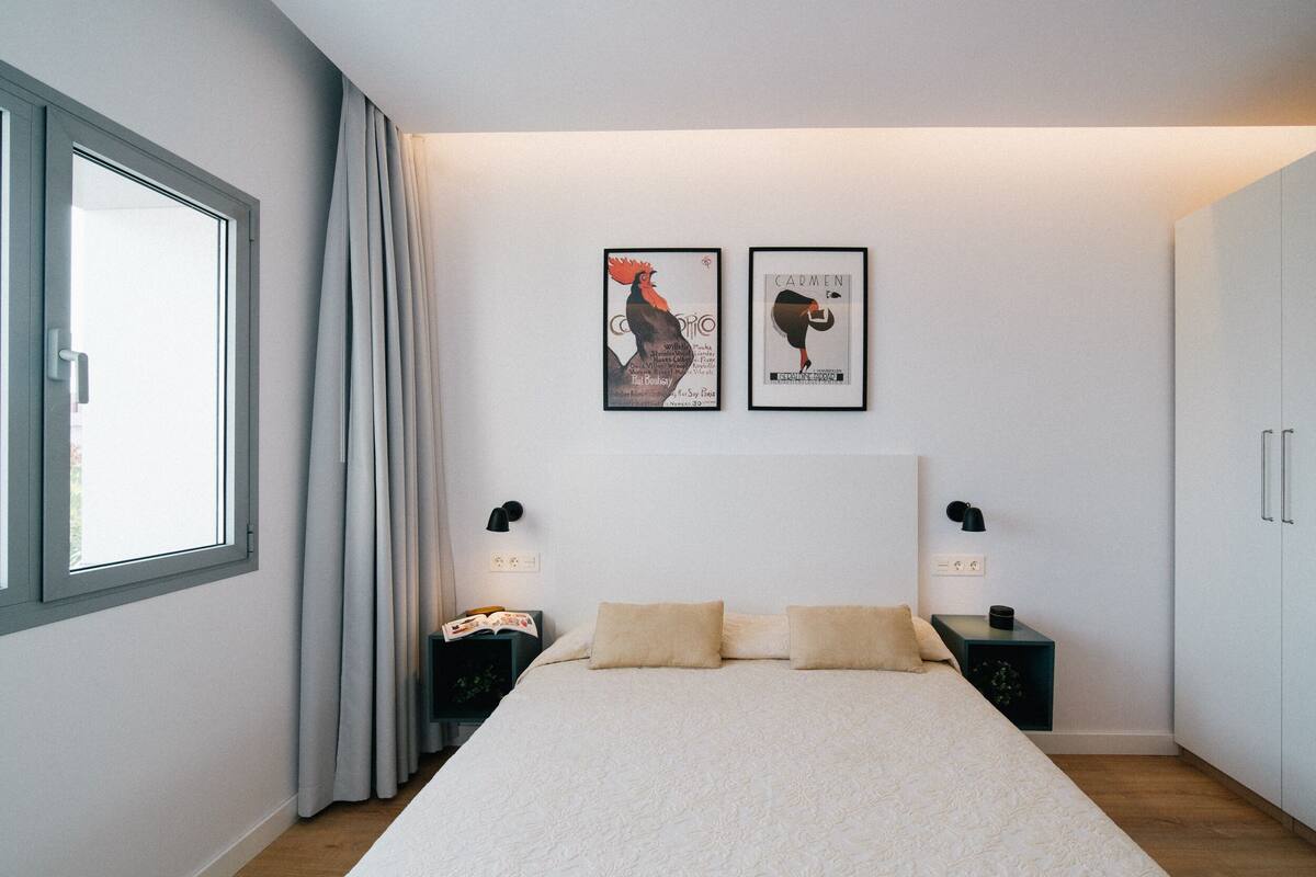 A modern bedroom with a large bed, two bedside tables, a window with curtains, and two framed artworks on the wall.