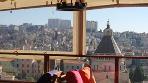 A room with a view of a cityscape, featuring a large window and a table with chairs. - Seedi Yousef Hostel & cafe (Nazareth)