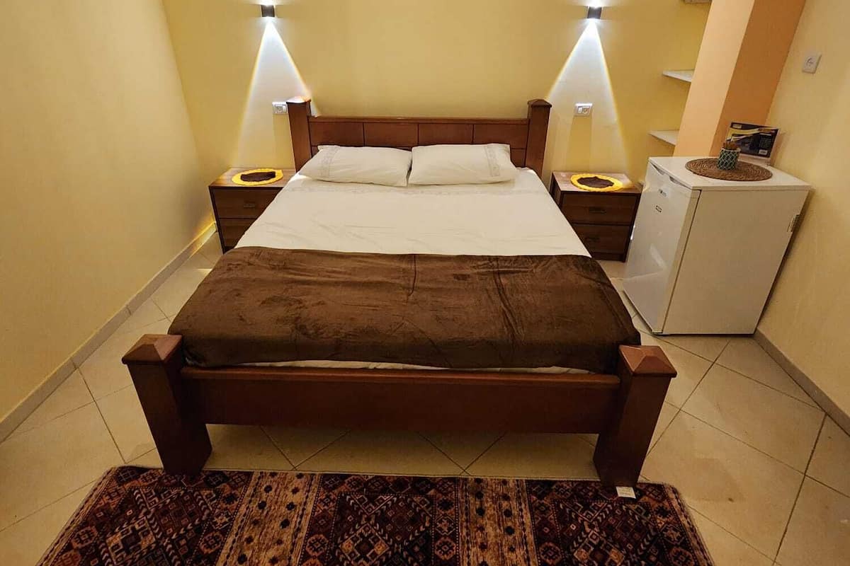 A bedroom with a wooden bed, two bedside tables, a nightstand, a small refrigerator, and a patterned rug.