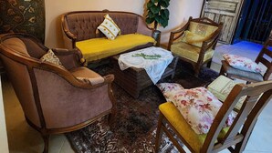 A cozy living room with a brown sofa, a yellow ottoman, and a floral armchair. A large green vase and a potted plant add greenery. The room has a tiled floor and a wooden door. - Seedi Yousef Hostel & cafe (Nazareth)