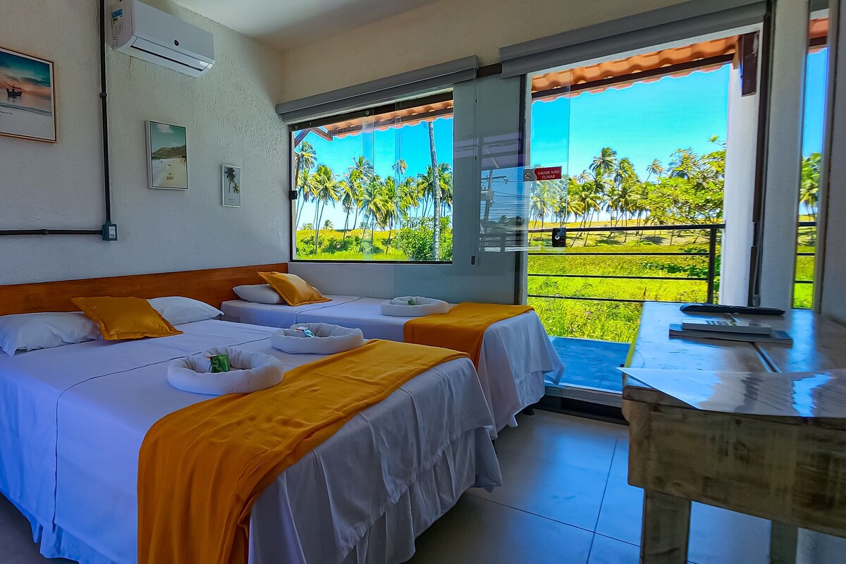 A room with two beds, a wooden door, and a balcony with a view of palm trees.