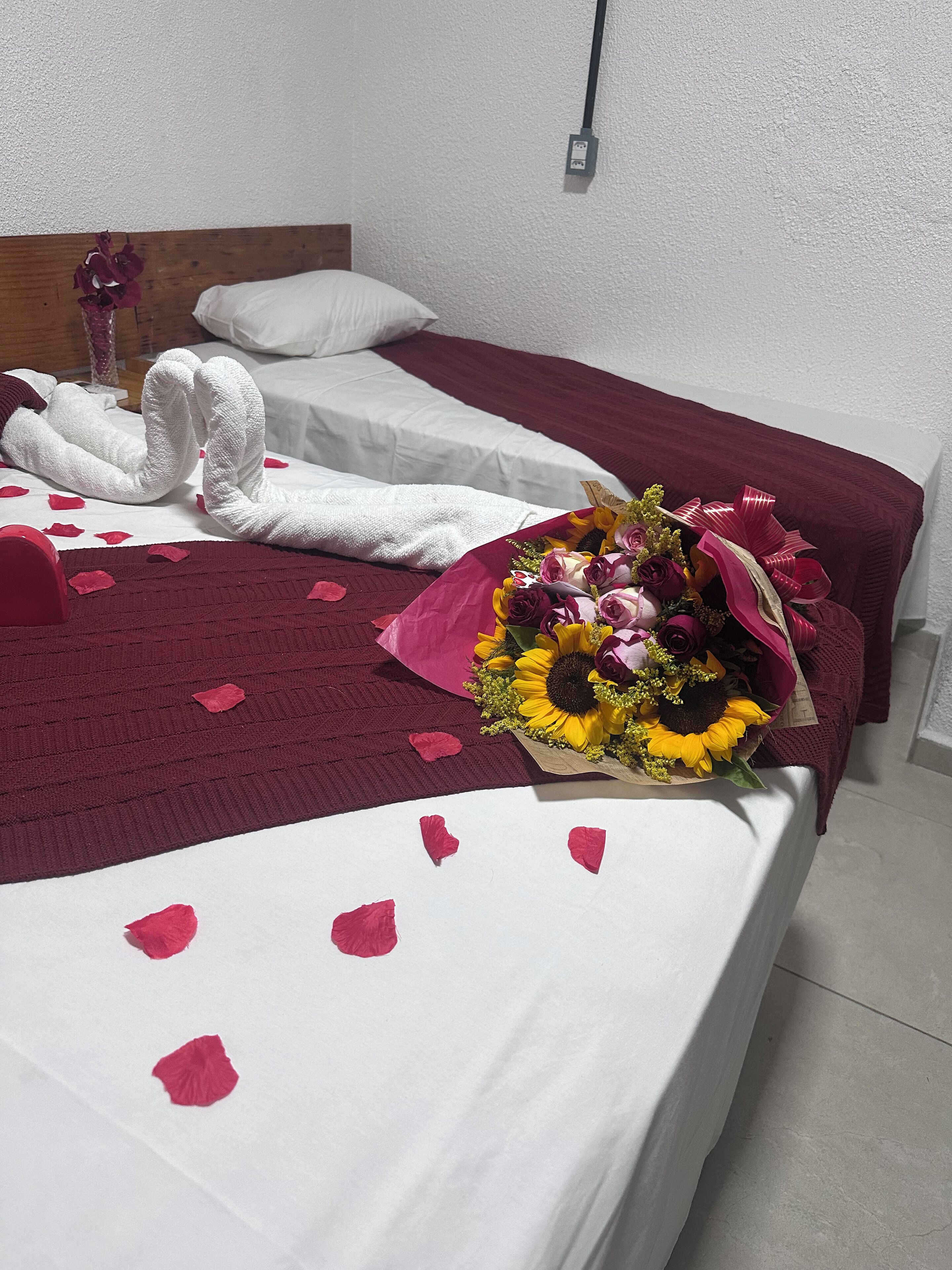 Una cama con una colcha blanca y burdeos, un ramo de flores y pétalos de rosa sobre la cama.