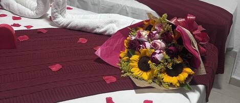 Una cama con una colcha blanca y burdeos, un ramo de flores y pétalos de rosa sobre la cama.