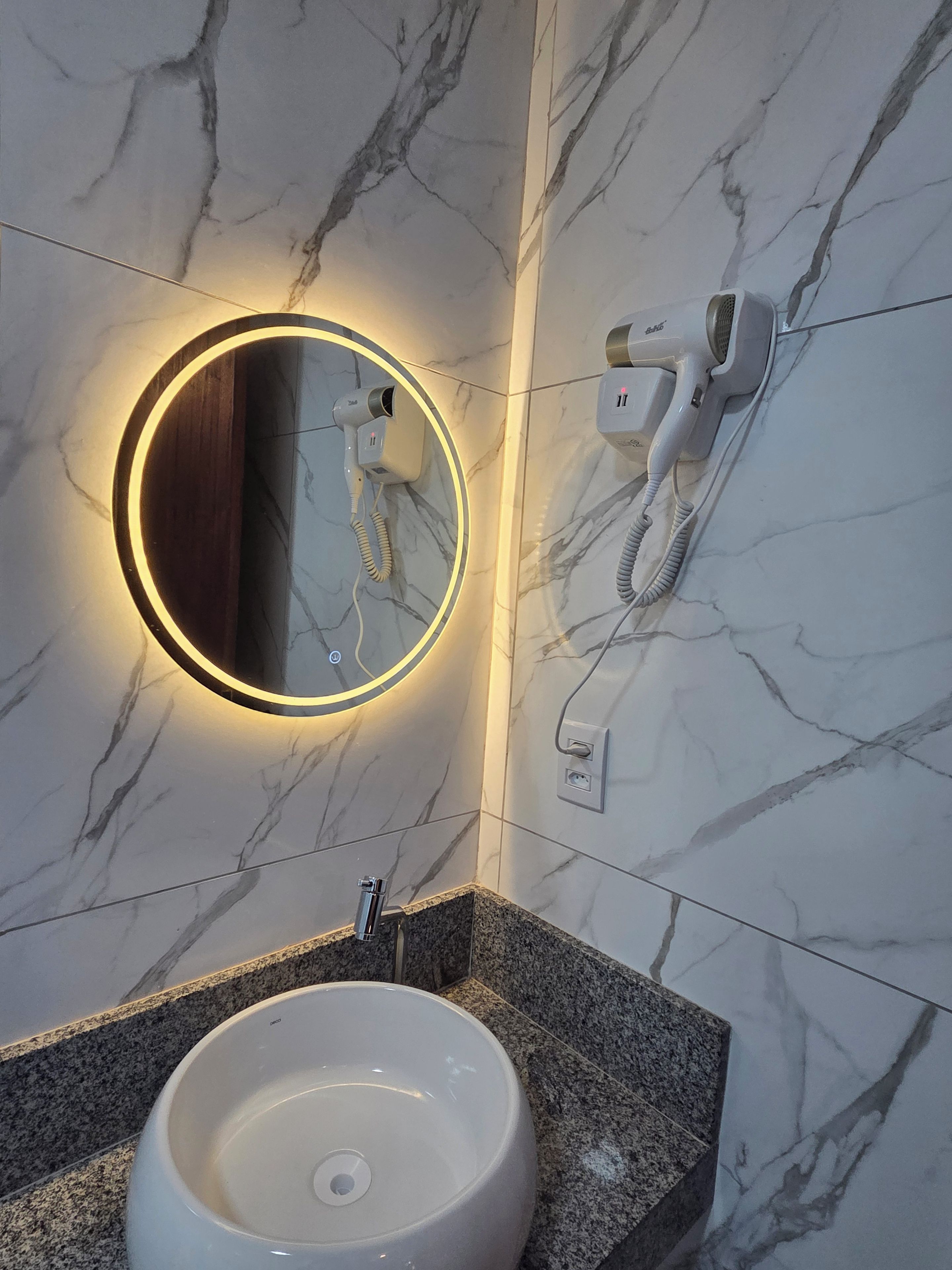A bathroom with a marble wall, a round mirror with a light, a hairdryer, and a sink with a granite countertop.