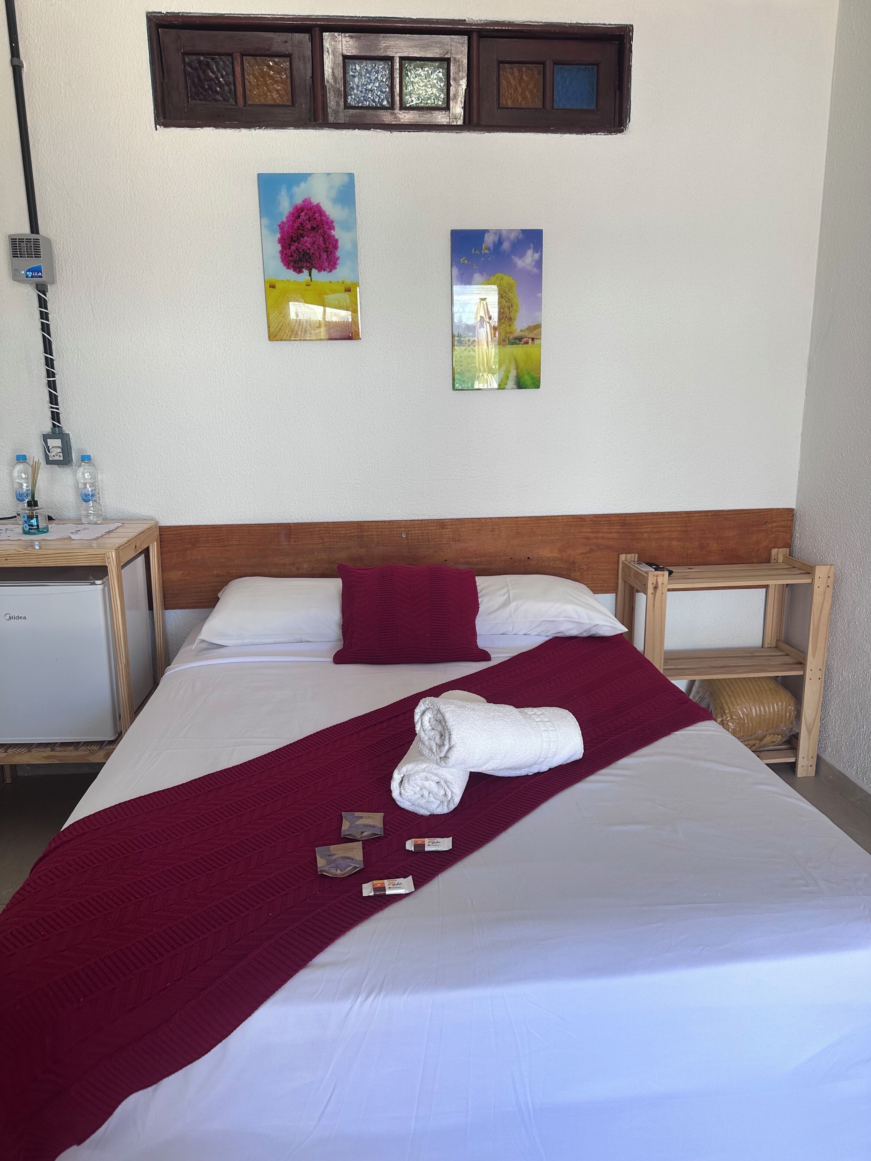A neatly made bed with a white and maroon bedspread, a rolled white towel, and two pillows. There are two bedside tables with lamps, a small refrigerator, and a wall-mounted power outlet. Two framed paintings are hung above the bed.