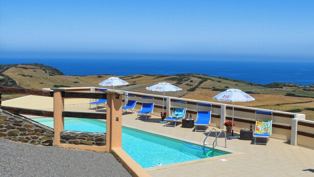 A pool area with blue lounge chairs and umbrellas, a stone wall, and a view of the ocean. - azienda agrituristica Finagliosu (Sassari)