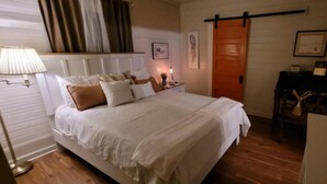 Ein Schlafzimmer mit einem Bett, Nachttischen, einer Lampe, Holzboden, einer Tür und einem kleinen Tisch mit einer Pflanze.