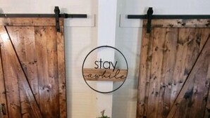 Ein Zimmer mit Holztüren, die sich verschieben lassen, einem kleinen schwarzen Tisch mit einem Topfpflanzen und einem runden Schild mit dem Schriftzug 'stay awhile'.