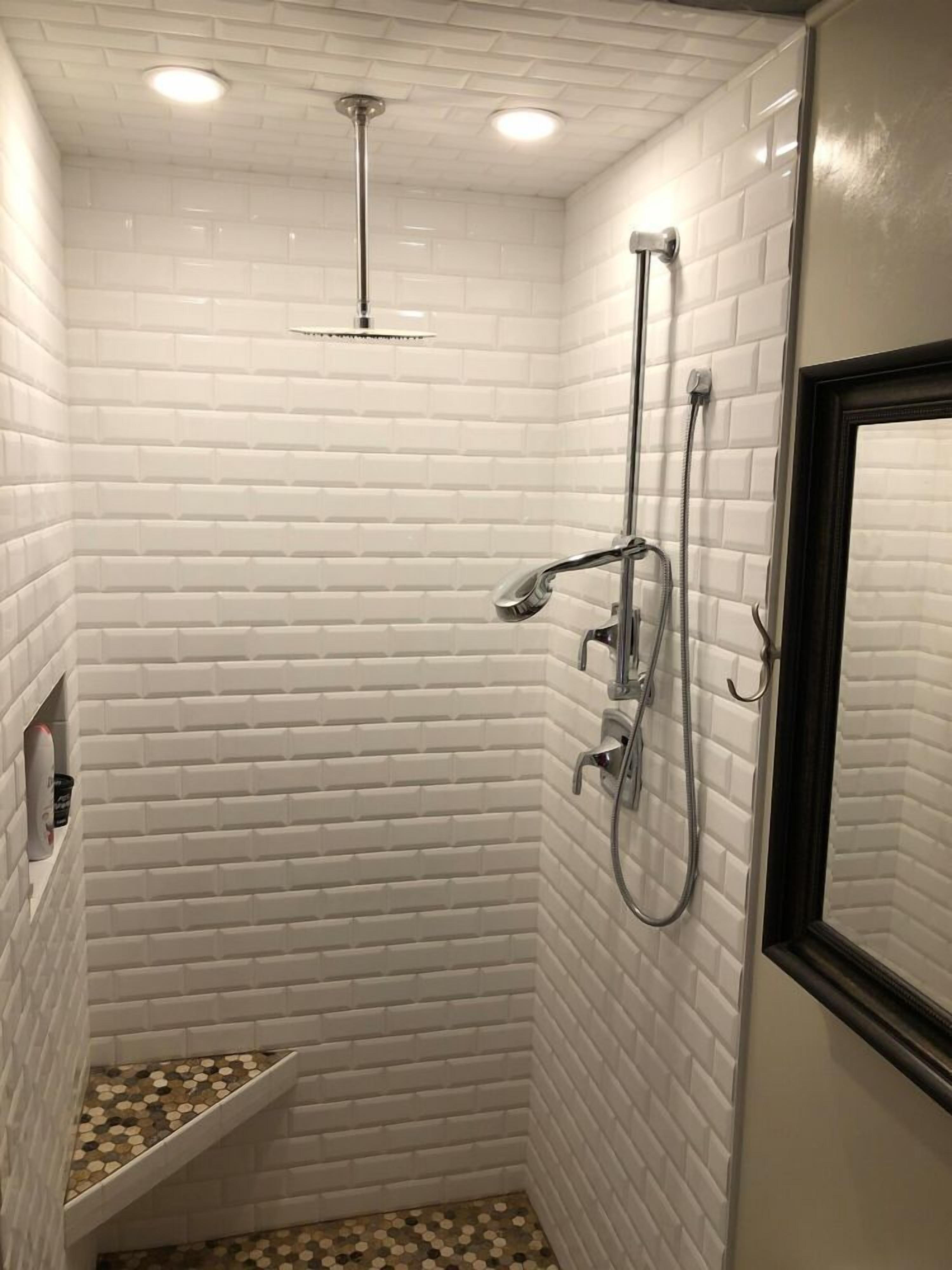 A small, tiled shower room with a white brick wall, a showerhead, a handheld shower, and a wall-mounted faucet.
