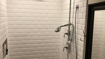 A small, tiled shower room with a white brick wall, a showerhead, a handheld shower, and a wall-mounted faucet.