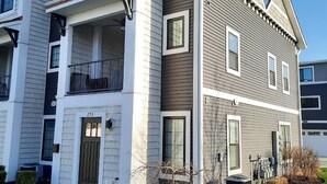 A multi-story residential building with a balcony on the second floor, a small front yard with a tree and shrubs, and a clear blue sky. - Spacious 3 Room Condo Near Michigan's Piers and Waterways (Grand Haven)