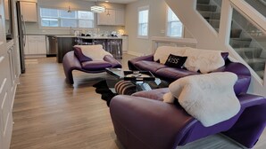 A modern living room with purple sofas and a glass coffee table, a kitchen in the background, and a staircase leading to an upper level. - Spacious 3 Room Condo Near Michigan's Piers and Waterways (Grand Haven)