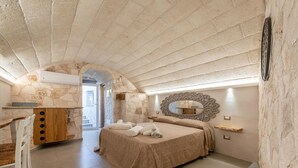 Quarto com parede de pedra, cama com colcha bege, cómoda de madeira e espelho com moldura decorativa.