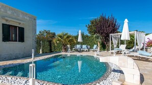 Piscina com borda azulejada, rodeada por espreguiçadeiras e guarda-sóis brancos, e um edifício com fachada de pedra e portadas.