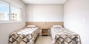 Habitación doble con dos camas, cabecero de madera, mesita de noche pequeña de madera y ventana con vistas a edificios.