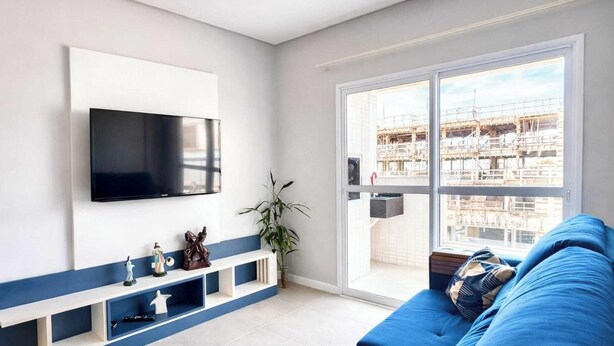 Ein modernes Wohnzimmer mit einem flachbildfernseher an der Wand, einem blauen Sofa, einem weißen Konsolentisch mit Dekoration und einer großen Schiebeglastür, die zu einem Balkon mit Blick auf eine Baustelle führt.