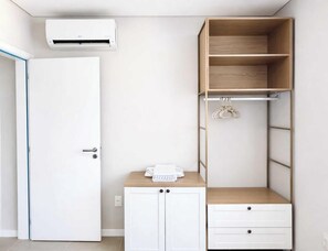Una habitación con una puerta blanca, un armario blanco con cajones, una estantería de madera con ropa colgada y un aire acondicionado instalado en la pared.