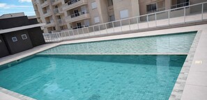 Una piscina con suelo de baldosas, barandilla blanca y un edificio con balcones al fondo.