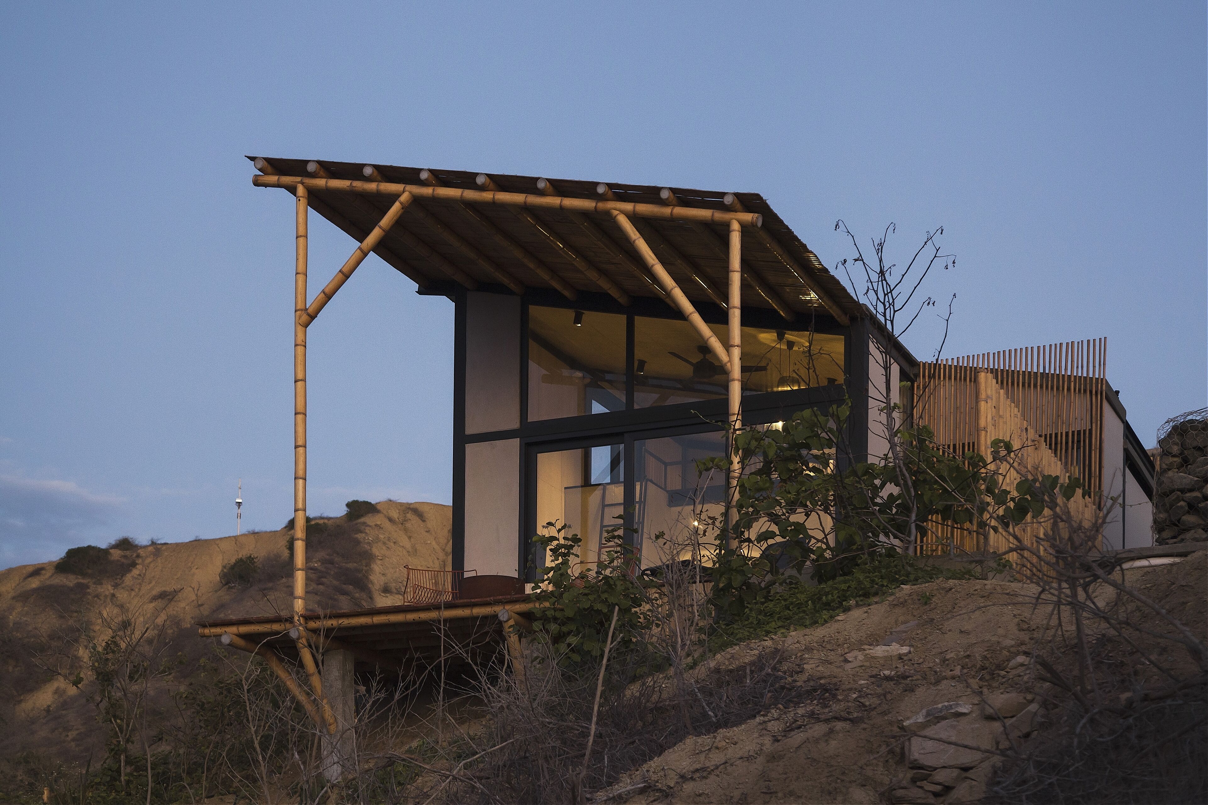 A modern, minimalist house with a metal framework and glass walls, situated on a hillside with a clear view of the surrounding landscape.