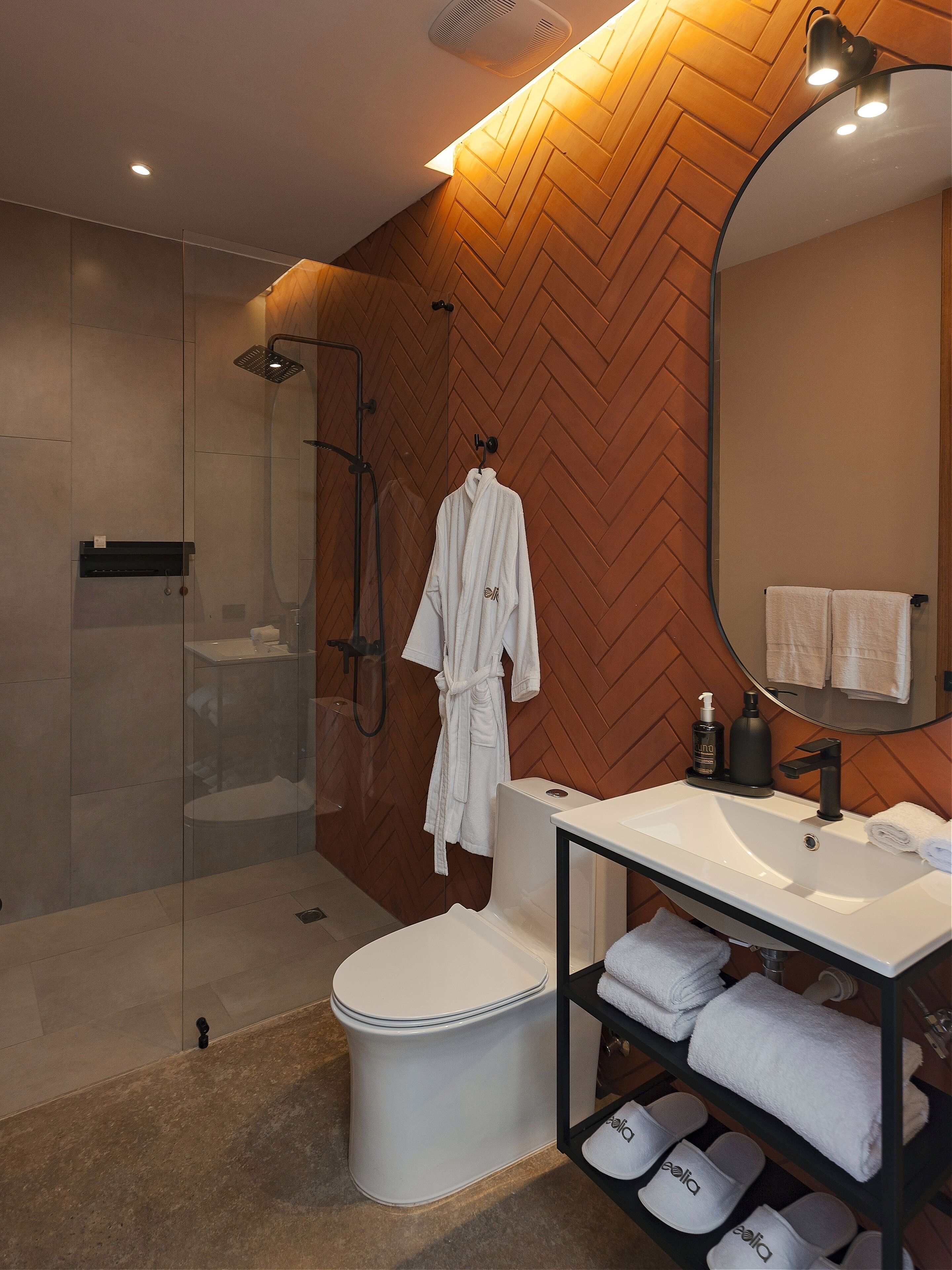 A modern bathroom with a glass-enclosed shower, a white toilet, a vanity with a mirror, and a shelf with towels and slippers.