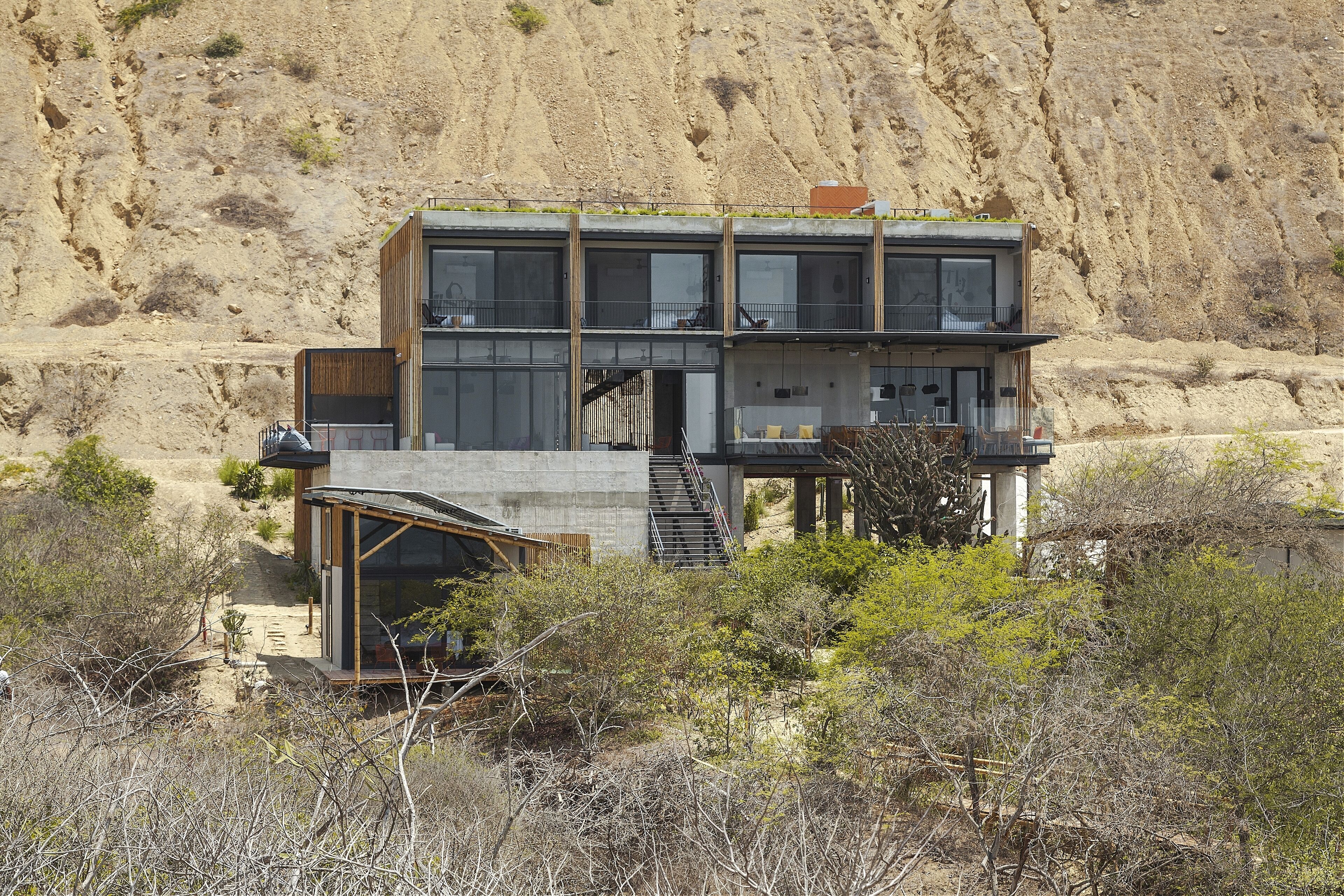 A modern two-story building with a staircase leading to the upper floor, surrounded by dry vegetation and a rocky hillside.