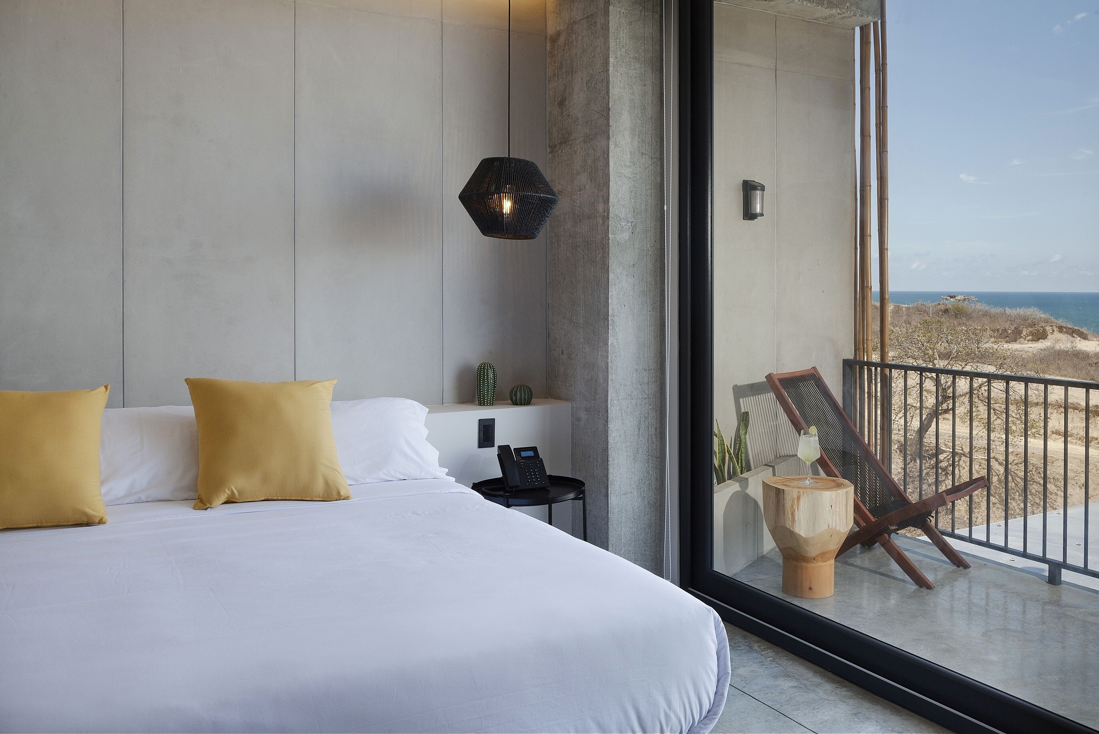 Habitación de hotel moderna con una cama grande, un teléfono en la mesita de noche y vista a la playa a través de un ventanal.