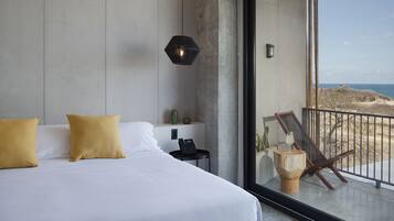 Habitación de hotel moderna con una cama grande, un teléfono en la mesita de noche y vista a la playa a través de un ventanal.