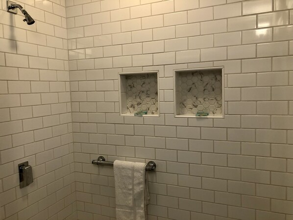 A bathroom with white subway tiles, a showerhead, two small shelves with toiletries, and a towel rack.