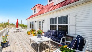 Een houten terras met een wit huis, een rood dak en tuinmeubels, waaronder een bank en stoelen.