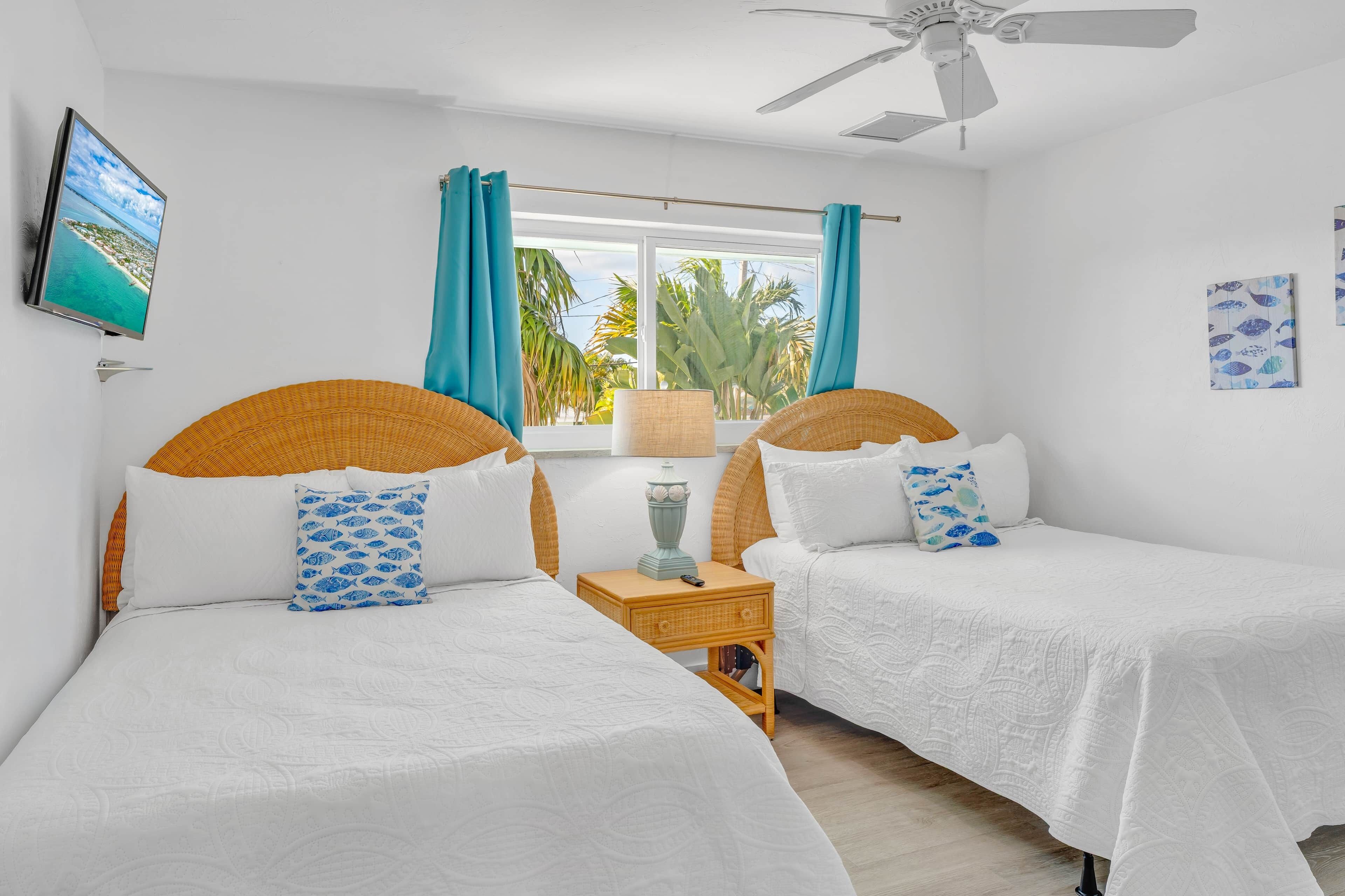 A hotel room with two beds, a ceiling fan, a window with curtains, a TV, and artwork on the walls.
