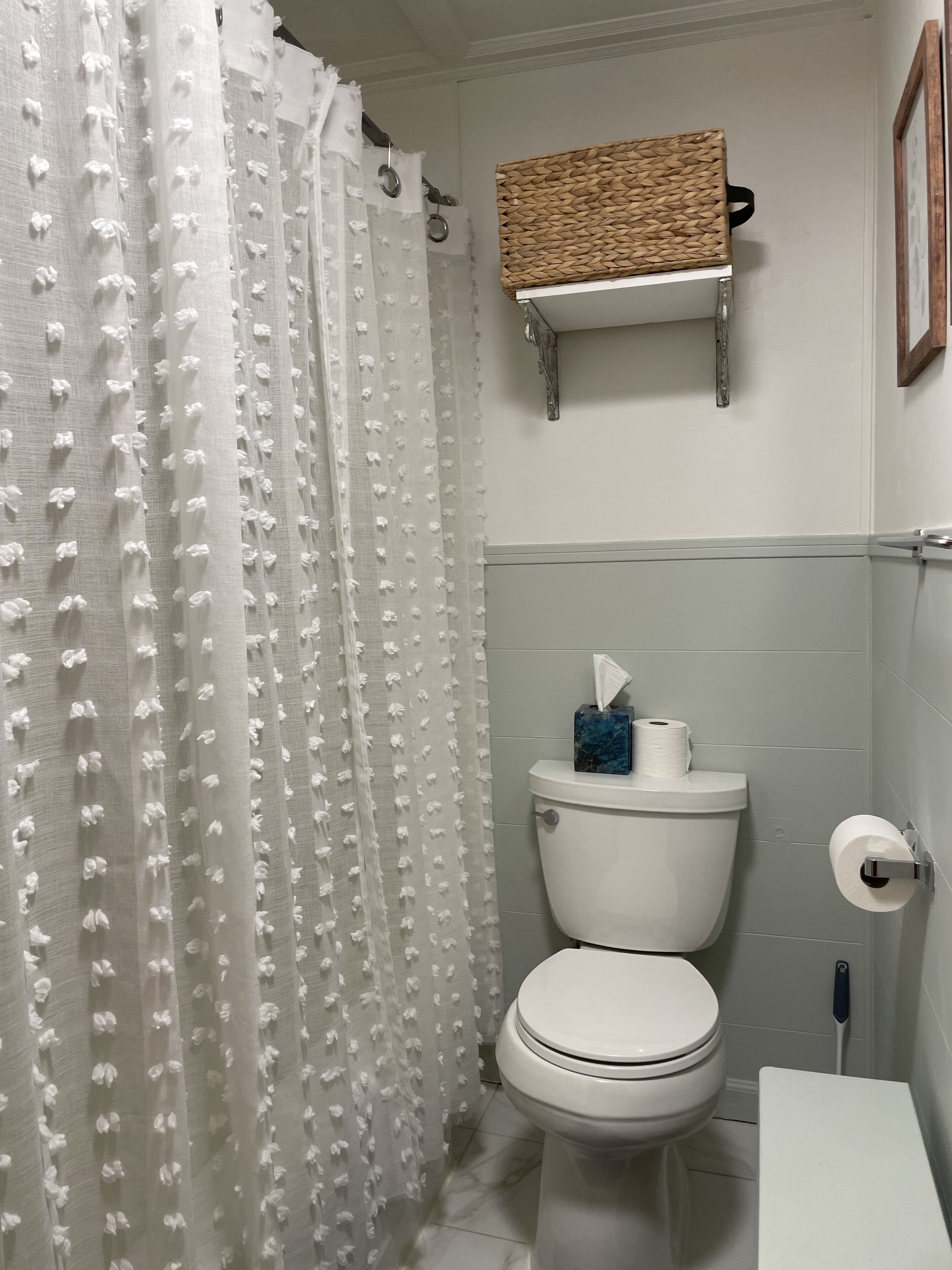 Un baño con inodoro, cortina de ducha, porta papel higiénico y repisa con cesta tejida.