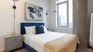 A modern hotel room with a large bed, a bedside table, a nightstand, a floor lamp, a wall-mounted artwork, and a window with a view of buildings. - 2BR Penthouse | Gym | Bellevue | Sophari Towers E (Bellevue)