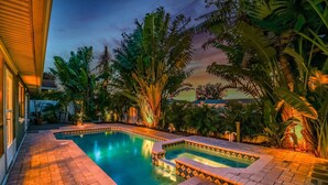 Una piscina con borde de baldosas, rodeada de plantas tropicales y un cielo al atardecer.