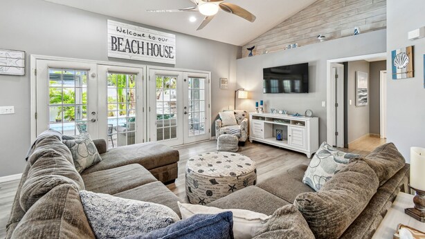 Un amplio salón con un gran sofá, un reposapiés redondo con motivos náuticos, un ventilador de techo y un cartel que dice "Welcome to our BEACH HOUSE".