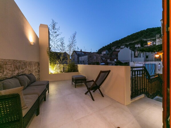 Un balcon avec un canapé, une chaise et une petite table. On aperçoit des maisons et des arbres au loin.