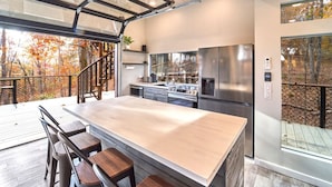 A modern kitchen with a large island, stainless steel appliances, and a view of autumn trees through large windows. - Skywalk Treehouse: 4 Treehouses Linked by Bridges (Sevierville)