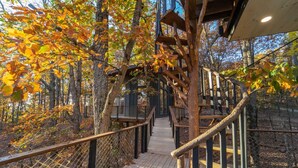 A wooden deck with a rope railing, surrounded by trees with autumn leaves. - Skywalk Treehouse: 4 Treehouses Linked by Bridges (Sevierville)