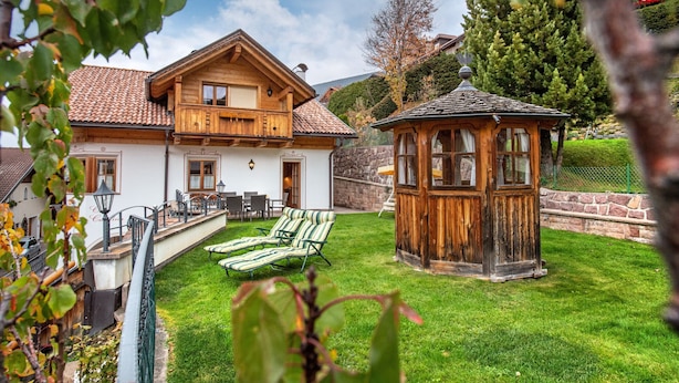 A wooden house with a balcony, a small wooden gazebo, and a grassy yard with lounge chairs. - Apartment Carolina 185A (Ortisei)