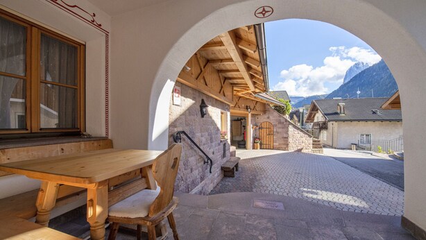 Ein Sitzbereich im Freien mit Holztisch und Bänken, ein Durchgang zu einer Bergblick und eine Steinsäule mit Laterne.