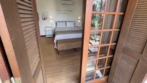 A bedroom with a wooden floor, a bed with striped bedding, a bedside table with a lamp, and a wall-mounted air conditioner. - Studios Aromas do Campeche - Floripa (Florianópolis)