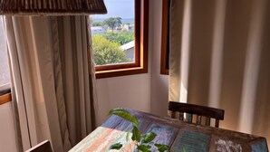 A room with a wooden table, a window with curtains, and a view of greenery and buildings outside. - Studios Aromas do Campeche - Floripa (Florianópolis)