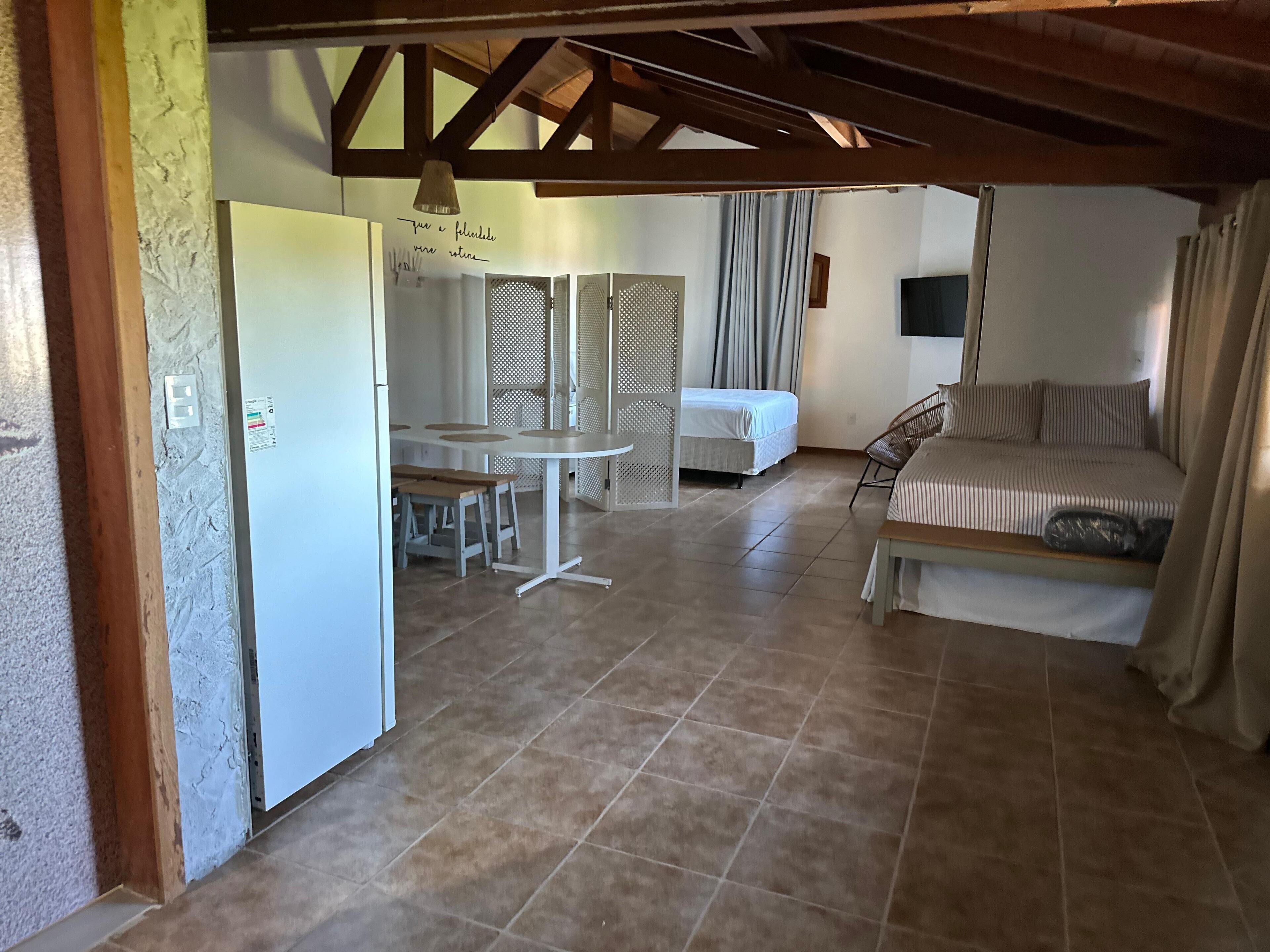 A room with a bed, a sofa, a dining table with benches, and a kitchen area.
