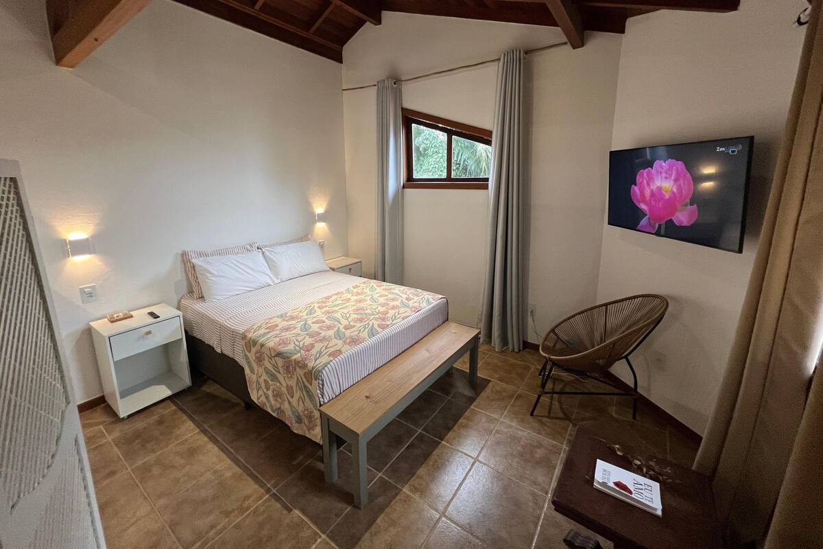 Une chambre à coucher comprenant un lit, une table de chevet, un banc, une chaise, un téléviseur et une fenêtre avec des rideaux.